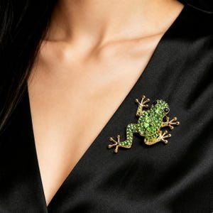Könnte beinhalten: Goldfarbene Froschbrosche mit grünen Kristallakzenten und einem schwarzen Auge. Die Brosche ist an einem schwarzen Kleidungsstück befestigt und zeigt die filigranen Details des Schmucks.