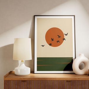 Op de afbeelding: Een ingelijste kunstdruk met een grote oranje zon tegen een crèmekleurige achtergrond, met een donkergroene horizonlijn en een zwerm zwarte vogels in de lucht. Het kunstwerk is ingelijst in een dunne zwarte lijst en toont een zonsondergang.