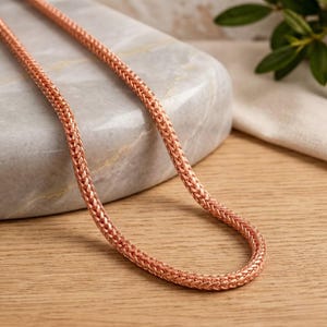 Könnte beinhalten: Eine roségoldfarbene Kettenhalskette mit einem dichten, geflochtenen Weizen- oder Fuchsschwanzmuster. Die Metallkette hat eine polierte Oberfläche und eine abgerundete, flexible Struktur, die ein durchgehendes, komplexes Flechtmuster aufweist.