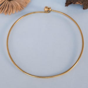 Könnte beinhalten: Ein dünner, kreisförmiger Choker aus goldfarbenem Metalldraht mit glatter, polierter Oberfläche. Die Halskette ist als durchgehender, offener Reif gestaltet und verfügt über einen kleinen, runden, kugelförmigen Verschlussmechanismus an der Oberseite.