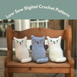 May include: Three crocheted cat toys sitting on a wooden bench. The cats are white, light blue, and beige. Each cat has embroidered eyes and a nose.