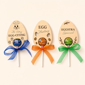 May include: Three Easter-themed lollipop favors. Each lollipop is attached to a wooden egg-shaped cutout with a colorful bow. Text on the cutouts reads: "It's Very Egg-Citing", "Egg Hunting", and "Eggstra Special".