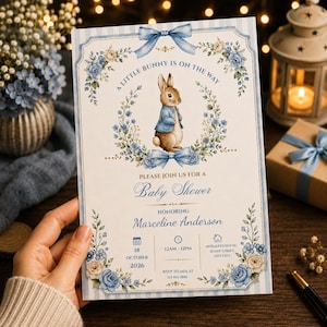 Puede incluir: Una tarjeta de invitación rectangular para baby shower con una ilustración en acuarela de un conejo marrón con chaqueta azul, rodeado de coronas de flores azules y crema, lazos azules y el texto A LITTLE BUNNY IS ON THE WAY y Baby Shower.