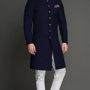 Könnte beinhalten: Eine marineblaue, langärmelige Sherwani-Jacke mit Stehkragen und einer Reihe metallischer Knöpfe auf der Vorderseite, kombiniert mit einer weißen Hose und schwarzen Slippern. Ein kleines, bunt gemustertes Einstecktuch steckt in der Brusttasche.