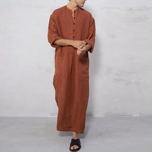 May include: A long-sleeved, floor-length robe in a solid burnt orange color. The garment features a round neckline, a partial button-down placket with five visible buttons, and two large patch pockets on the front. The fabric has a textured appearance.