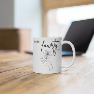 May include: A white ceramic mug featuring a black line art illustration of a hand holding a lily flower. The text "Hello" is printed in a simple font, while the word "fourty" is written in a cursive script with "est. 1984" below it.