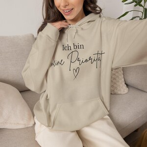 May include: A beige hooded sweatshirt featuring a front kangaroo pocket and a drawstring hood. The chest displays black text that reads Ich bin meine Priorität with a small hand-drawn heart symbol centered below the lettering.