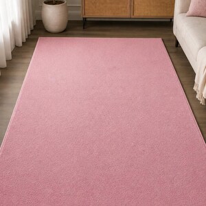 May include: A rectangular area rug in a solid, soft pink color with a low-pile, textured surface. The rug features finished, bound edges in a matching pink tone, providing a clean and uniform border around the entire perimeter of the piece.