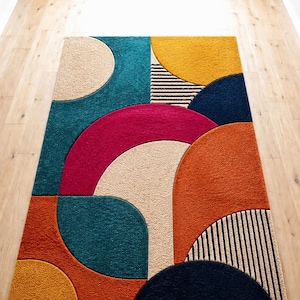 Op de afbeelding: Een rechthoekig vloerkleed met een geometrisch patroon van overlappende cirkels en halve cirkels in teal, mosterdgeel, magenta, oranje-rood en marineblauw, met secties van zwart-witte strepen en getextureerde beige accenten.
