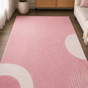 May include: A rectangular area rug featuring a solid pink background with a large white semi-circle on the bottom left corner, a white semi-circle on the middle right edge, and a series of thin, vertical white lines extending from the right side.