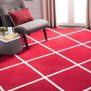 May include: A large rectangular area rug featuring a vibrant red background with a prominent white grid pattern of intersecting lines. The rug covers the floor space, extending beneath a gray upholstered chair with a red accent pillow.