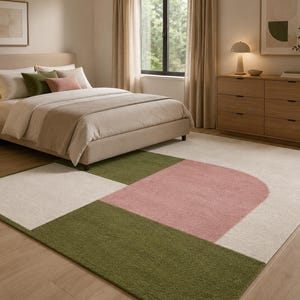 May include: A rectangular area rug featuring a geometric pattern with large blocks of cream, olive green, and a soft pink semi-circle shape. The rug has a low-pile texture and is positioned on a light wood floor in a bedroom setting.