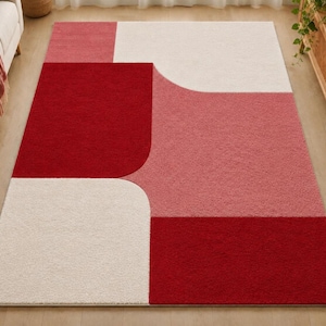 May include: A rectangular area rug featuring a geometric pattern of large, solid color blocks in deep red, soft pink, and off-white. The design consists of rounded corners and straight edges, creating a balanced composition of contrasting hues.