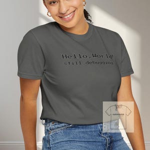 May include: A gray t-shirt with the text "Hello, World! still debugging" in a simple, thin font. The shirt is worn with blue jeans. The model is smiling and sitting on a black stool.