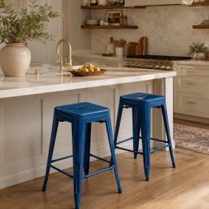 May include: Two blue metal bar stools with square seats and footrests are positioned in a kitchen setting. The stools have a glossy finish and a minimalist design. The kitchen features a white countertop, wooden shelves, and a neutral color palette.