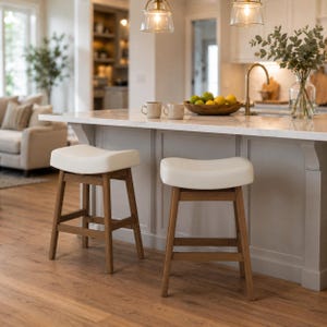 May include: Two wooden bar stools with cream-colored seats are positioned in front of a kitchen island. The stools have a light brown wooden frame and a curved seat. The kitchen features a marble countertop and pendant lights.