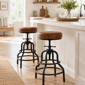May include: Two industrial-style bar stools with brown leather seats and black metal frames. The seats have a textured appearance. The stools are adjustable and have a footrest. The stools are in a kitchen setting.