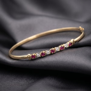 Puede incluir: Una pulsera rígida de oro con bisagra que presenta una hilera de rubíes rojos y diamantes blancos redondos alternados, engastados en una banda de metal pulido. La pulsera cuenta con un mecanismo de cierre seguro y un perfil redondeado.