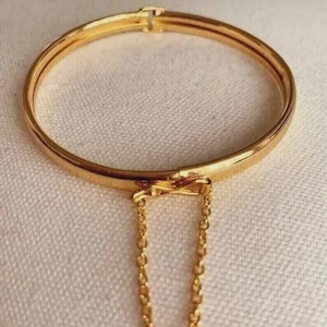 May include: A gold-toned metal bangle bracelet featuring a smooth, circular band with a hinged clasp mechanism at the top. A delicate, thin gold-toned chain is attached to the front of the bangle, forming a small, dangling loop below the band.
