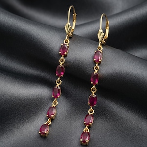 May include: A pair of long drop earrings featuring five oval-cut deep red rubies set in yellow gold. Each earring is suspended from a gold lever-back closure, with the rubies arranged in a vertical line connected by small gold jump rings.