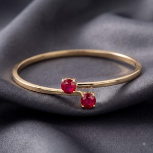 May include: A gold bypass bangle bracelet featuring two round, faceted red rubies set in four-prong settings at the open ends. The polished gold band has a smooth, slender profile that curves to showcase the vibrant, circular gemstones.