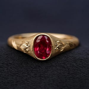 Könnte beinhalten: Ein goldener Ring mit einem zentralen, oval geschliffenen roten Edelstein. Die Ringschiene hat eine texturierte, matte Oberfläche und ist auf beiden Seiten der Fassung mit einem Blumenmotiv graviert, das ein detailliertes botanisches Design zeigt.