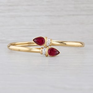 May include: A gold bypass bangle bracelet featuring two pear-shaped red gemstones at the open ends. Each gemstone is accented by a small silver-toned band set with tiny clear stones, creating a polished look with a smooth, rounded gold band.
