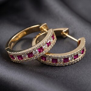 Op de afbeelding: Een paar gouden oorringen met een centrale rij vierkant geslepen rode robijnen, geflankeerd door twee rijen kleine, ronde, helderwitte diamanten. De oorbellen hebben een scharnierende sluiting en een gepolijste gouden afwerking over het geheel.