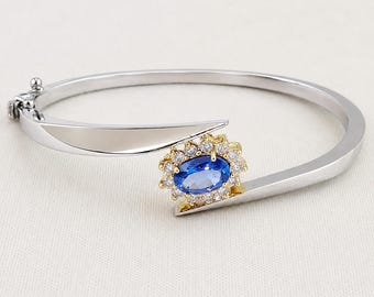 May include: A silver-toned hinged bangle bracelet featuring a bypass design. One end terminates in a polished silver point, while the other end showcases an oval blue gemstone surrounded by a halo of small clear stones set in gold-toned metal.