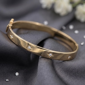May include: A gold bangle bracelet with star-shaped accents, each set with a small, sparkling diamond. The bracelet is displayed on a gray, textured surface, with additional loose diamonds scattered around.