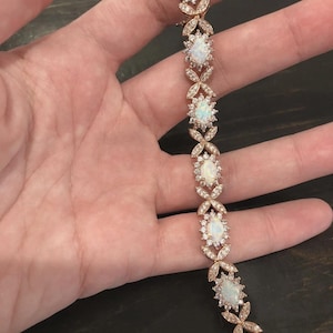 May include: A rose gold tone bracelet featuring a series of oval-shaped iridescent opals, each surrounded by a halo of small clear stones. The links between the opal settings are shaped like small, delicate leaves or petals, creating a continuous chain.
