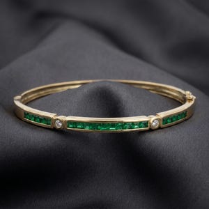 May include: A gold hinged bangle bracelet featuring a channel setting of square-cut green gemstones. The design includes two small, round, clear stones set in circular gold bezels that divide the green gemstone sections along the band.