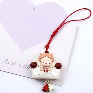 Op de afbeelding: Een witte bedel in de vorm van een envelop met een slapend poppenontwerp. De bedel heeft een rood touwtje en twee kleine belletjes die aan de onderkant hangen. De bedel staat op een witte achtergrond met een hartvormige kaart.