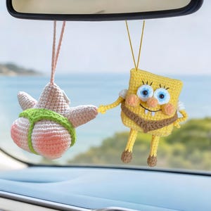 May include: Two crocheted hanging ornaments featuring a yellow rectangular character with blue eyes and a pink star-shaped character wearing a green thong. Both figures are suspended by thin strings from a car rearview mirror.