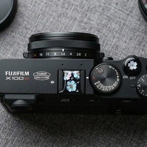 Op de afbeelding: Een zwarte Fujifilm X100VI digitale camera met een bloemmotief op de flitsschoenafdekking en de ontspanknop. De bovenkant toont de merknaam, het model, het lenssysteem en ronde draaiknoppen voor sluitertijd en belichting.