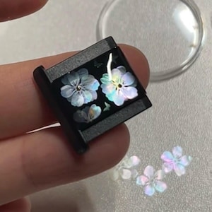 May include: A square black charm featuring an iridescent floral inlay with three distinct flower shapes that display a pearlescent sheen. The surface is smooth and dark, contrasting with the light-reflecting petals of the floral design.
