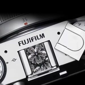 May include: A silver-coloured metal hot shoe cover for a Fujifilm camera, featuring an embossed floral design with five petals. The cover is inserted into the camera's hot shoe slot, positioned near the Fujifilm logo and a 35mm lens marking.