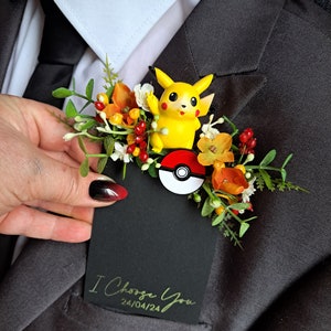 Może przedstawiać: Czarna kartka z złotym napisem "I Choose You" i datą "24/04/24". Mała żółta figurka Pikachu z czerwoną i białą Pokéballą jest przymocowana do bukietu pomarańczowych i białych kwiatów.