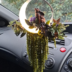 May include: A car charm with a brown rabbit figurine inside a glowing crescent moon. The moon is surrounded by greenery, purple crystals, and dark berries. The charm hangs from a string and is suspended in front of a car dashboard.