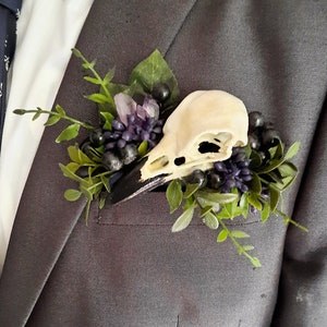 May include: A small white bird skull with a black beak is surrounded by purple and green flowers and berries. The skull is pinned to a gray suit jacket.