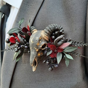 May include: A black and gold ram skull with celestial details is surrounded by burgundy and black flowers. The skull is a decorative element for a wedding or special occasion.