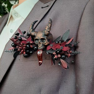 May include: A bronze skull with horns and a red crystal pendant is attached to a lapel pin. The pin is surrounded by black berries, red leaves, and dark red floral accents. The pin is attached to a brown suit jacket.