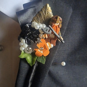 Op de afbeelding: Een corsage met een plastic dwergfiguurtje dat een grijze bijl vasthoudt, versierd met witte kunstparels, een goudkleurig blad, een oranje bloem met een strass-steentje, zilveren sterretjes en een groen stoffen blad op een zwart lint.