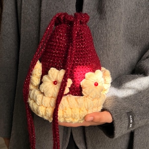 May include: A drawstring pouch crafted from deep red, glittery yarn, featuring a base and decorative trim in cream-coloured yarn. The pouch is adorned with three-dimensional cream flowers, each centred with a small, shiny red bead.