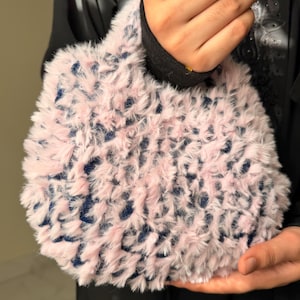 Puede incluir: Un bolso pequeño con un exterior de piel sintética de color rosa claro y textura esponjosa, con una base de tela azul oscuro visible a través del pelaje. El bolso tiene una forma redondeada con un asa integrada y un aspecto suave y peludo.