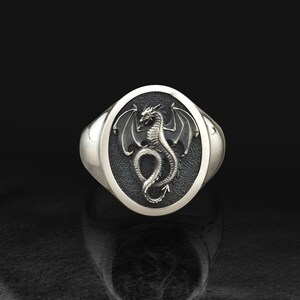 Puede incluir: Un anillo de sello plateado con una cara ovalada que presenta un motivo de dragón detallado en relieve. El dragón se muestra con las alas extendidas y la cola enroscada sobre un fondo oscuro y texturizado que contrasta con la banda de metal pulido.