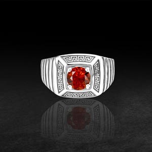 Könnte beinhalten: Ein silberfarbener Ring mit einem runden, leuchtend roten Edelstein in der Mitte. Die quadratische Vorderseite des Rings ist mit einem detaillierten griechischen Mäandermuster verziert, während die Ringschiene vertikale Rillen aufweist.