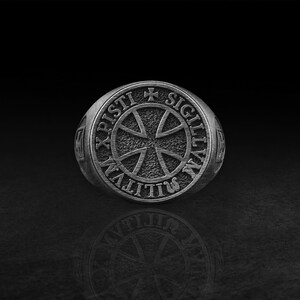 Könnte beinhalten: Ein silberfarbener Metallring mit einer kreisförmigen Vorderseite und strukturiertem Hintergrund. Die Mitte zeigt ein Kreuzsymbol, umgeben von der Inschrift SIGILLVM MILITVM XPISTI, mit einem kleinen Kreuz oben im Kreis.