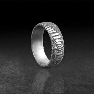 May include: A silver-toned ring featuring a textured exterior surface that mimics the appearance of reptile scales. The band has a smooth, brushed metallic interior finish and a rounded profile, showcasing a detailed, repeating pattern across the outer circumference.