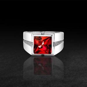 Könnte beinhalten: Ein silberfarbener Ring mit einem großen, quadratisch geschliffenen, leuchtend roten Edelstein in der Mitte. Die Ringschiene hat eine glatte, polierte Oberfläche mit texturierten, mattierten Seitenteilen, die sich nach hinten verjüngen.